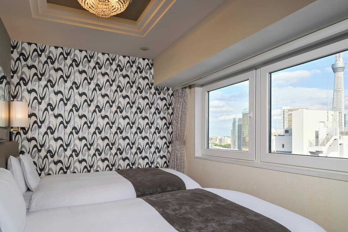 A hotel room with a patterned wallpaper, a large bed with white and brown bedding, a chandelier, and a window with a cityscape view.
