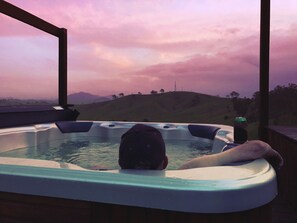 A hot tub with a person reclining inside, overlooking a hilly landscape during sunset. - Dam It Getaway- Homestead (Bowman Farm)