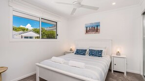 Une chambre à coucher avec un lit, deux tables de chevet, un ventilateur de plafond, une fenêtre donnant sur une maison et un tableau accroché au mur.