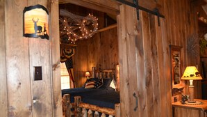 A rustic wooden room with a bed, a nightstand, and a wall-mounted lamp. - The Guest House (Schellsburg)