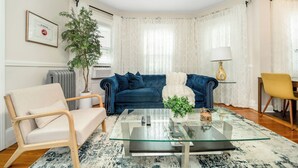 Una sala de estar con un sofá azul, una mesa de centro de vidrio, un sillón beige, una planta en maceta y una lámpara.