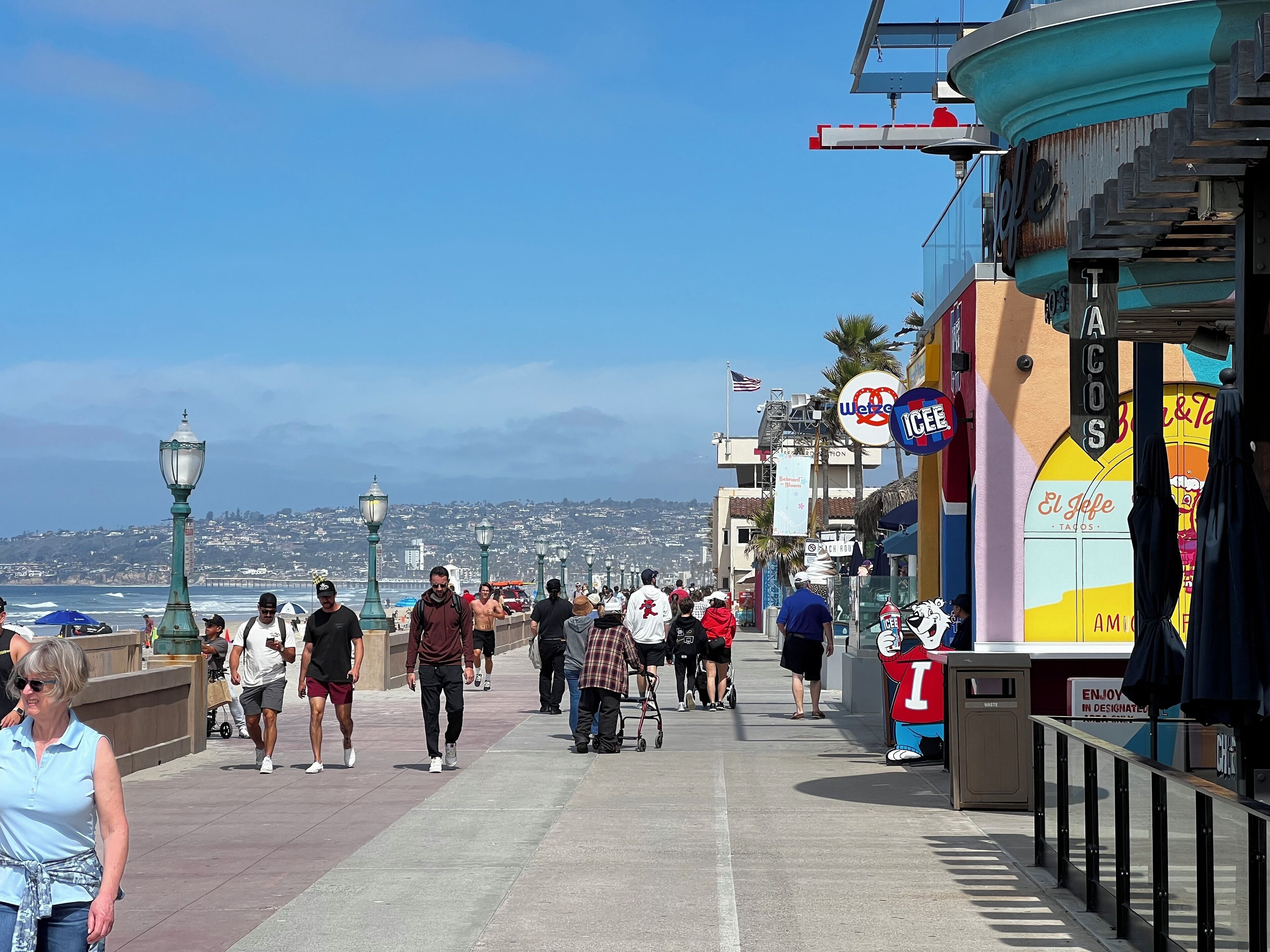 Une promenade animée où l’on peut voir des gens se promener, avec une vue sur le front de mer et divers commerces, dont un restaurant de tacos.
