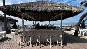 A beachside bar with a thatched roof, white plastic chairs, and a view of the ocean.