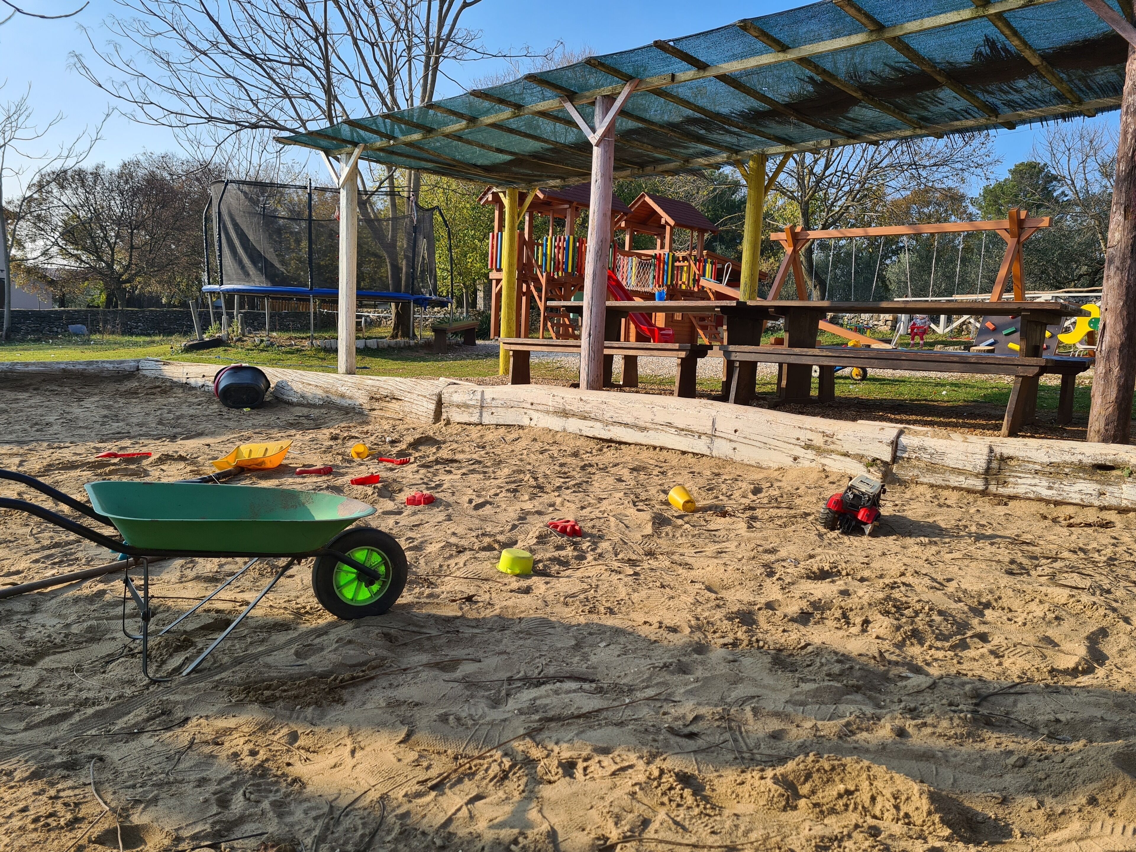 Un parque infantil con zona de juegos de arena, un conjunto de columpios de madera y una zona de estar cubierta.