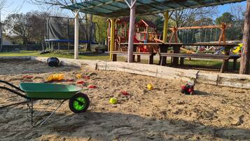 Un parque infantil con zona de juegos de arena, un conjunto de columpios de madera y una zona de estar cubierta.
