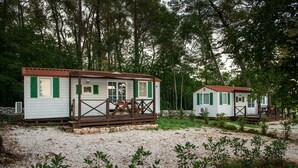 Two small, white cabins with green shutters, surrounded by trees and gravel paths.