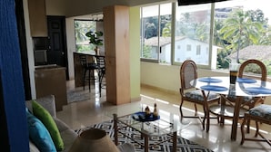 A spacious living area with a dining table, chairs, and a large window offering a view of palm trees and buildings. - Newly Listed Amazing, Serene resort with Beautiful Sunsets & Blue Waters  (PUERTO VALLARTA)