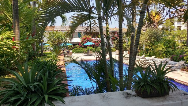 A pool area with palm trees, a tiled deck, and a building with a tiled roof in the background. - Newly Listed Amazing, Serene resort with Beautiful Sunsets & Blue Waters  (PUERTO VALLARTA)