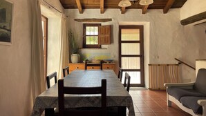 Un comedor rústico con una mesa de madera, sillas y un banco. Se ve una planta en maceta y una ventana con cortinas.