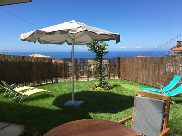 Ein Garten mit einem Holztisch, zwei Liegestühlen, einem kleinen Baum und einem großen Sonnenschirm.