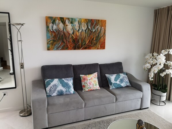 Un sofá gris con tres cojines decorativos, una lámpara de pie, un cuadro de flores y una orquídea en maceta.