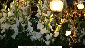 Eine Statue einer religiösen Figur mit Krone, umgeben von weißen Blumen und flackernden Lichtern.
