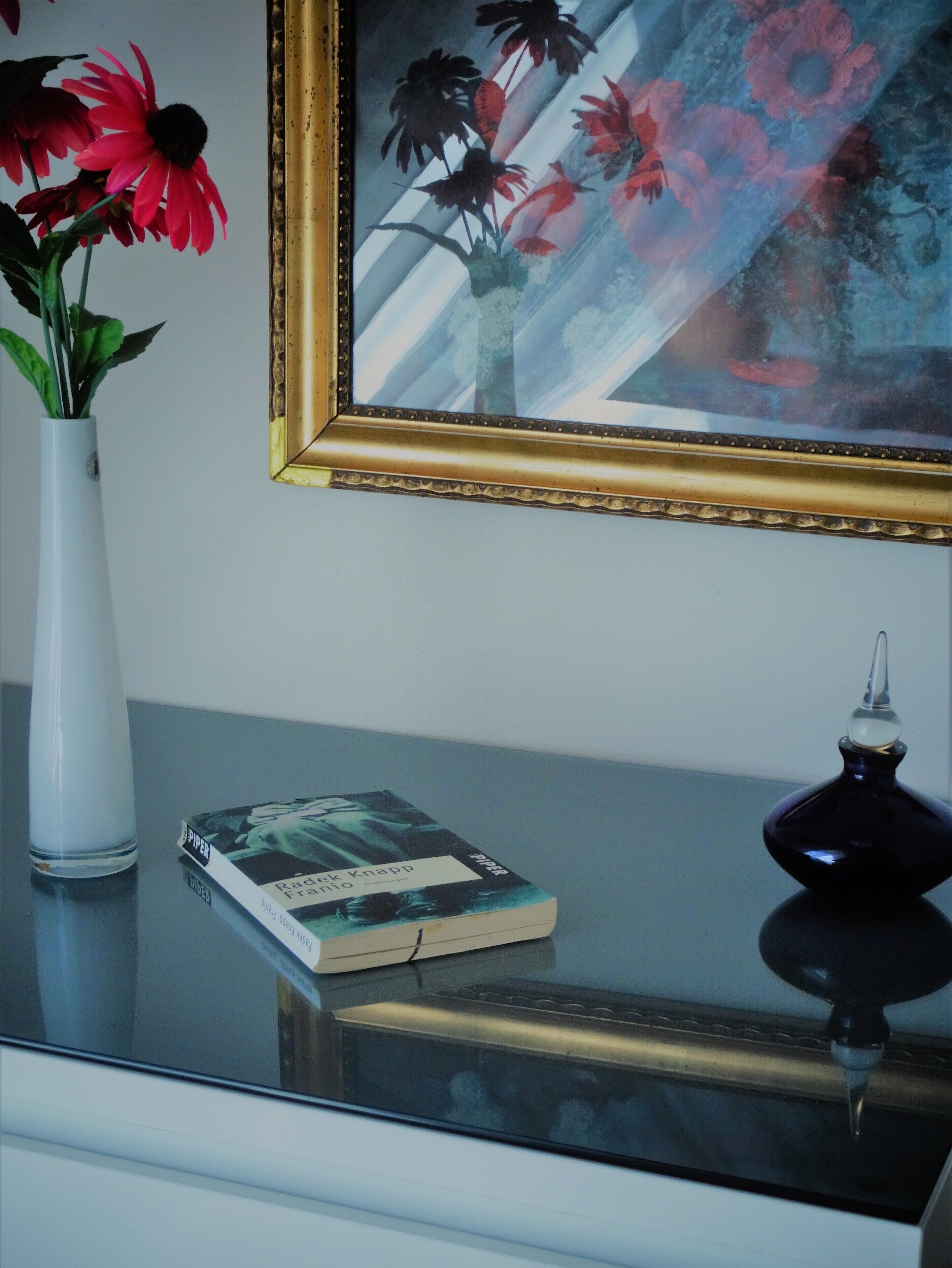 Une table en verre sur laquelle se trouvent un livre, un vase avec des fleurs et une bouteille décorative.