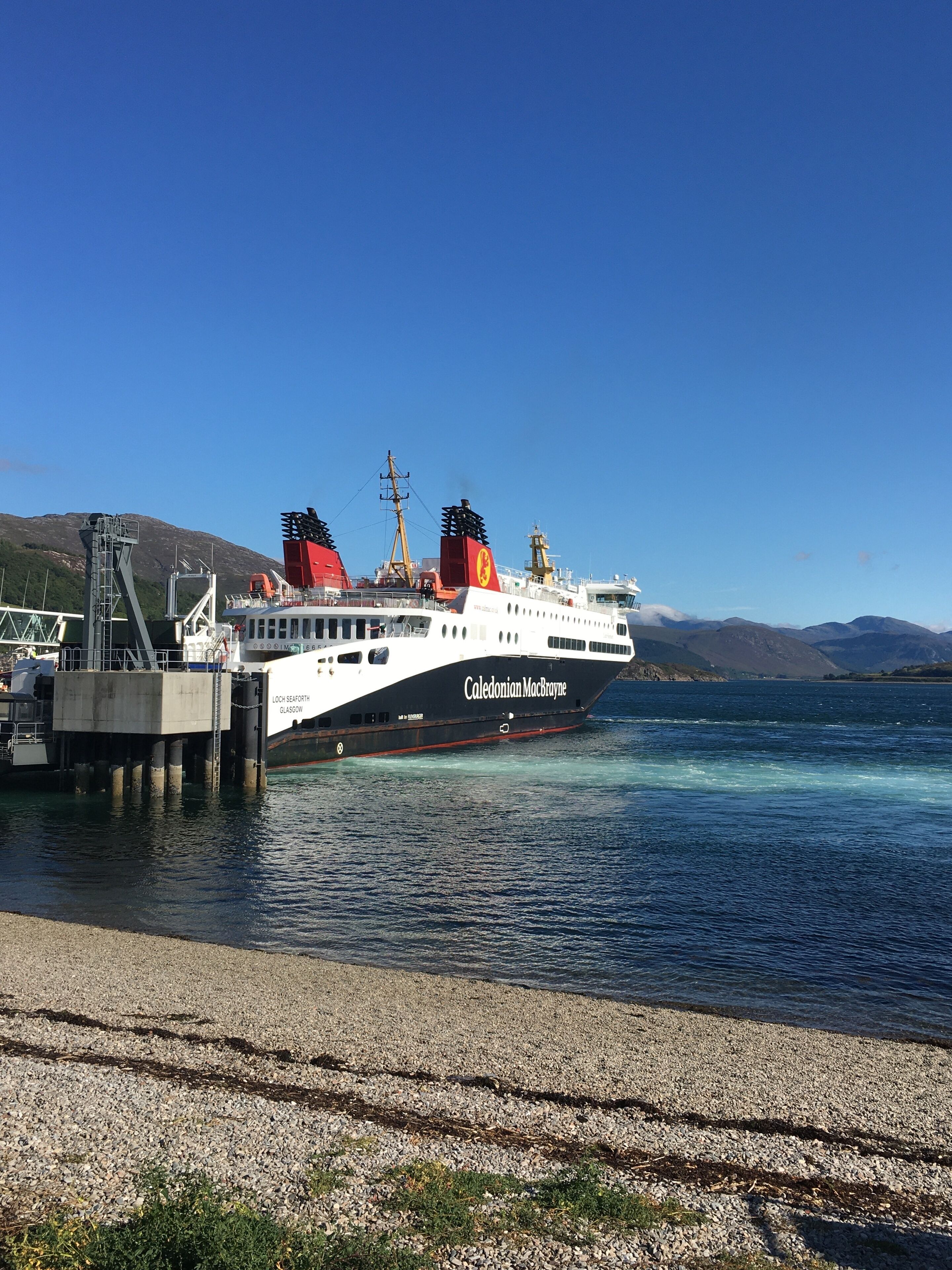 Een veerboot met de naam 'Caledonian MacBride' ligt aangemeerd aan een pier, met een heldere lucht en bergen op de achtergrond.
