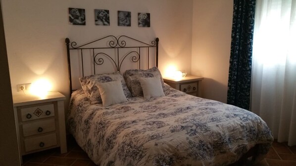 Une chambre à coucher comprenant un lit, deux tables de chevet, une commode, une lampe fixée au mur et une fenêtre avec des rideaux.