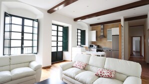 Un salón espacioso con un sofá y sillón blancos, una mesa de centro oscura, y una cocina al fondo.