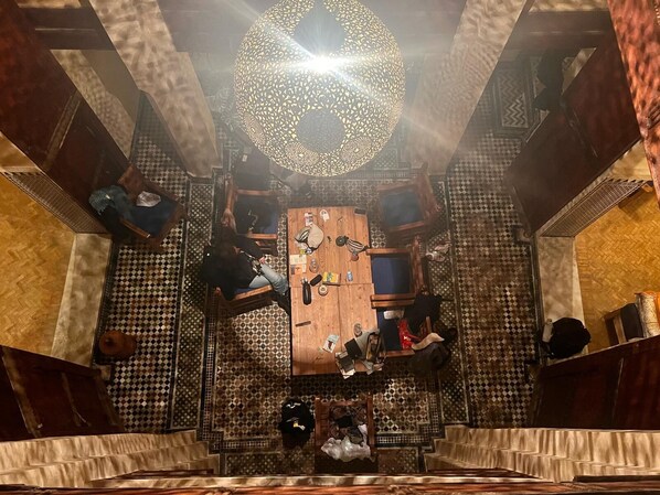 A room with a central wooden table surrounded by chairs, a patterned floor, and a decorative ceiling light.