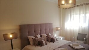 A neatly made bed with a large headboard, two bedside tables with lamps, a chair, and a window with curtains. - Beautiful new two bedroom apartment in Albufeira, Algarve<br> (Albufeira)