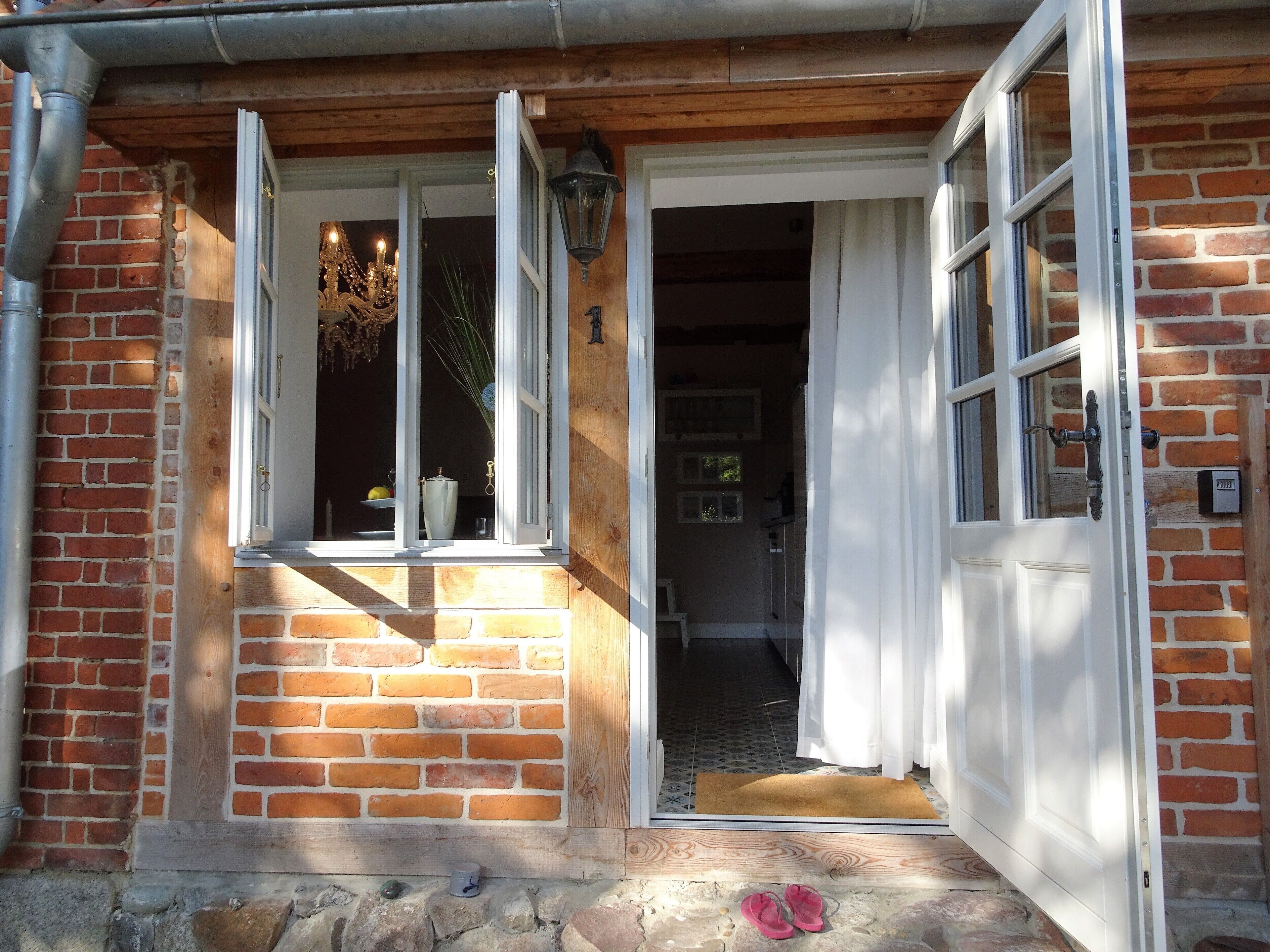 A brick house with a wooden porch, a white door, and a window with a curtain.