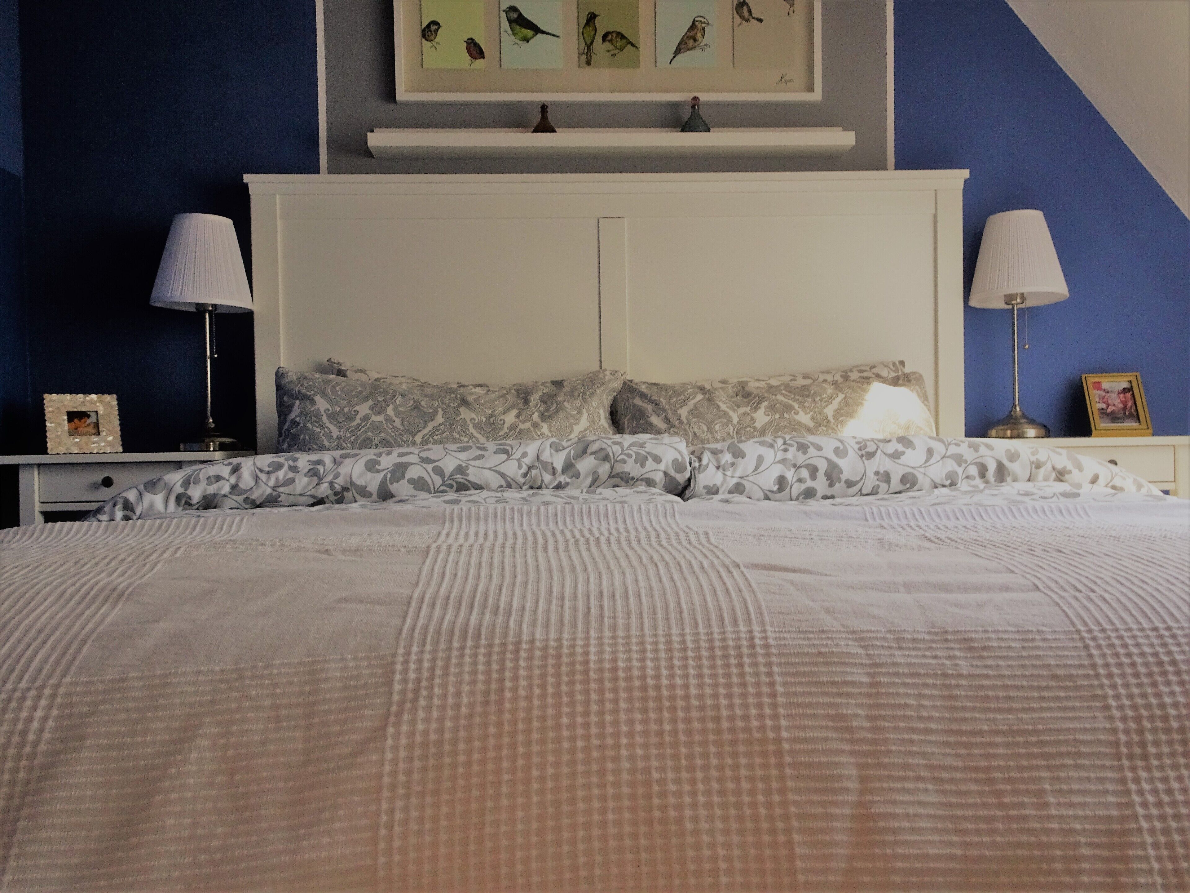 Un dormitorio con una cama, dos mesitas de noche, una mesita auxiliar, un estante con marcos y una lámpara en cada mesita de noche.