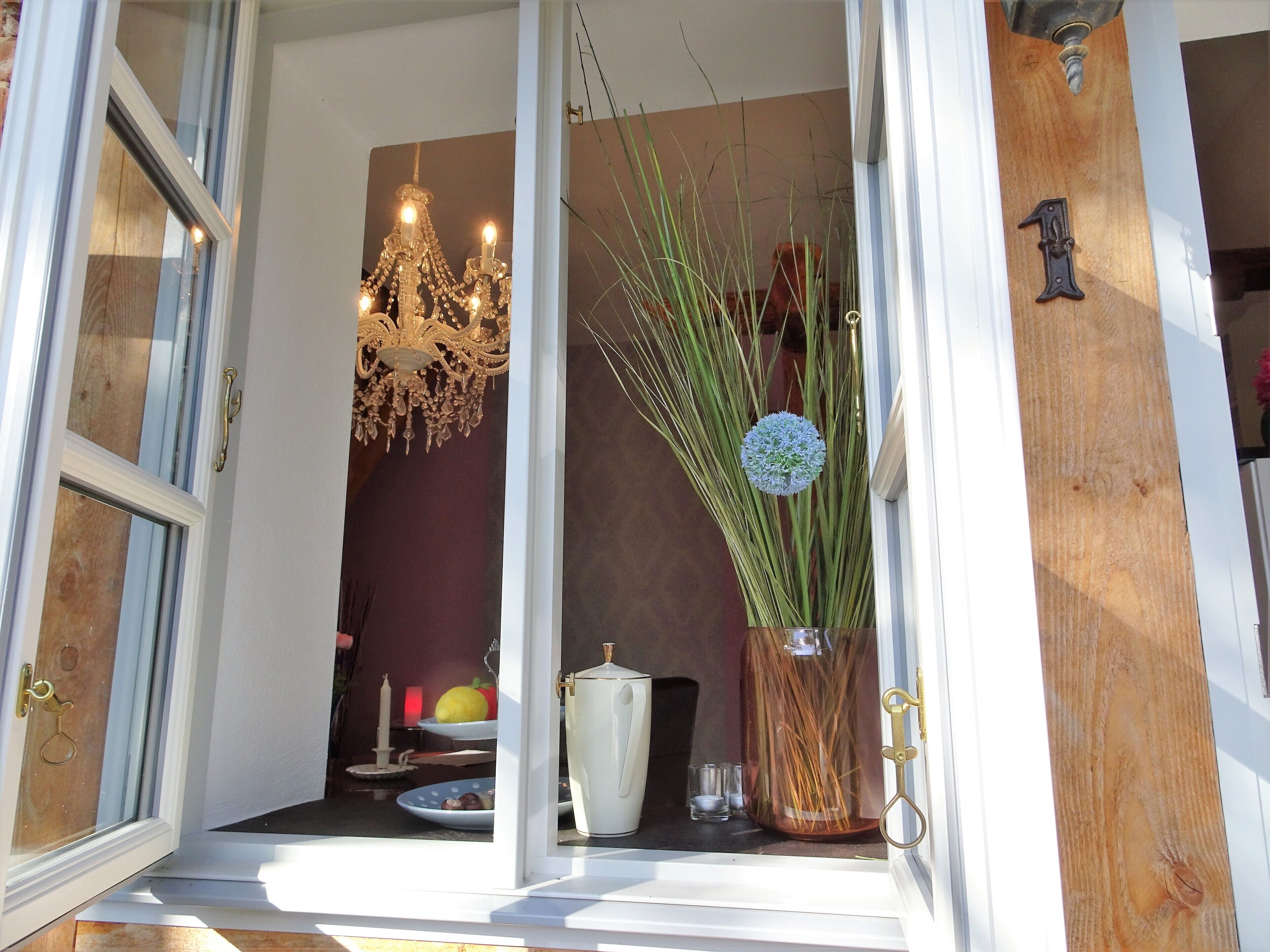 An open door leading to a room with a chandelier, a vase with tall plants, and a wooden door with a brass handle.