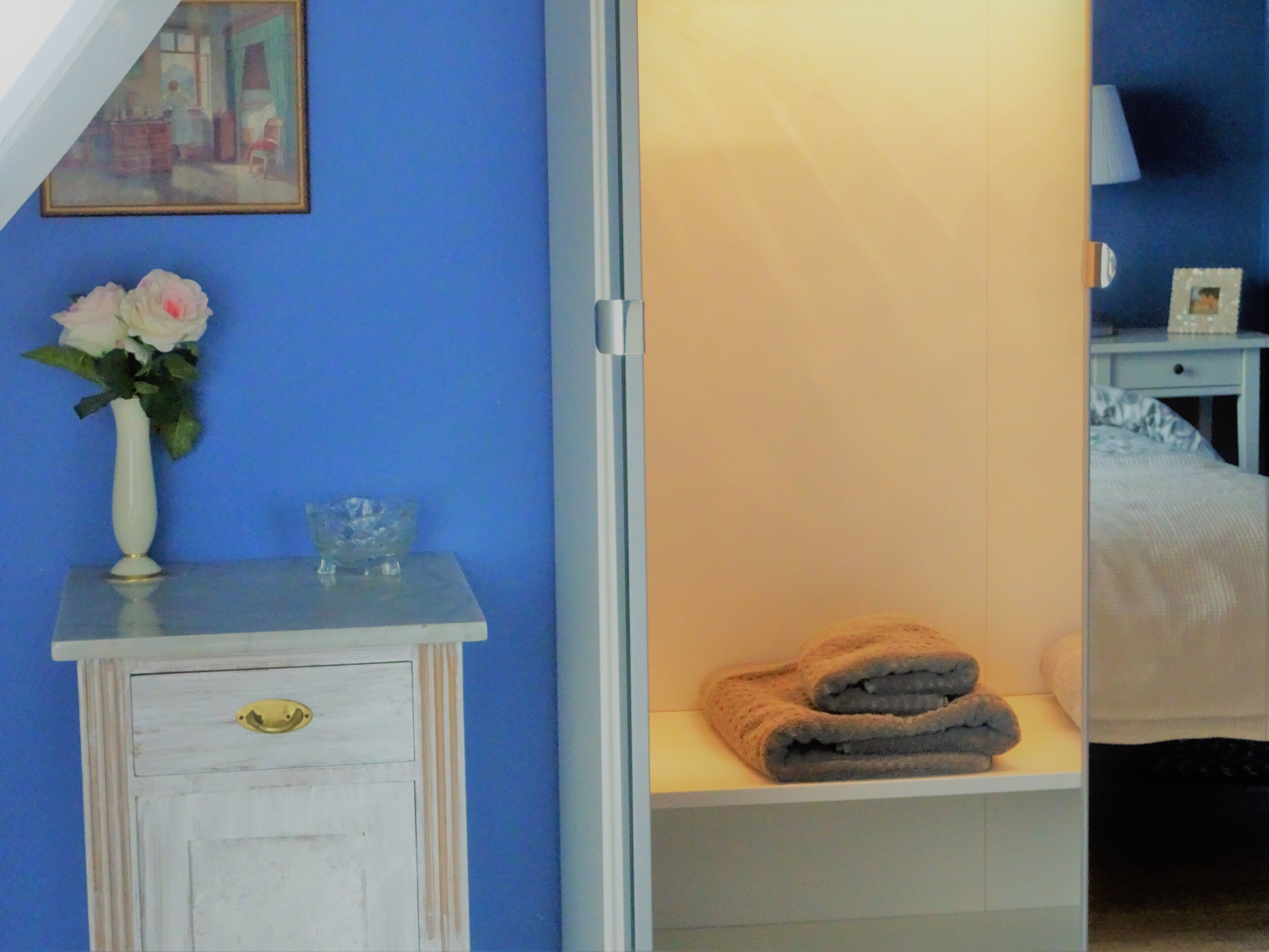 Quarto com parede azul, porta branca, mesa pequena com um vaso de flores, prateleira com toalhas dobradas e um quadro na parede.