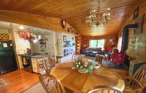 A cozy wooden dining area with a round table and chairs, a chandelier, and a kitchen visible in the background.