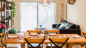 Un comedor con una mesa de madera dispuesta para cuatro personas, rodeada de sillas. Hay una estantería a la izquierda y un sofá a la derecha.