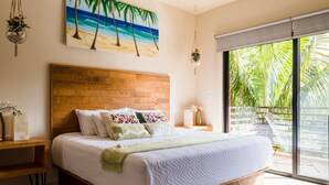 Un dormitorio con cabecera de madera, cama con ropa de cama blanca, un cuadro de una playa y vistas a palmeras a través de un ventanal corredero.