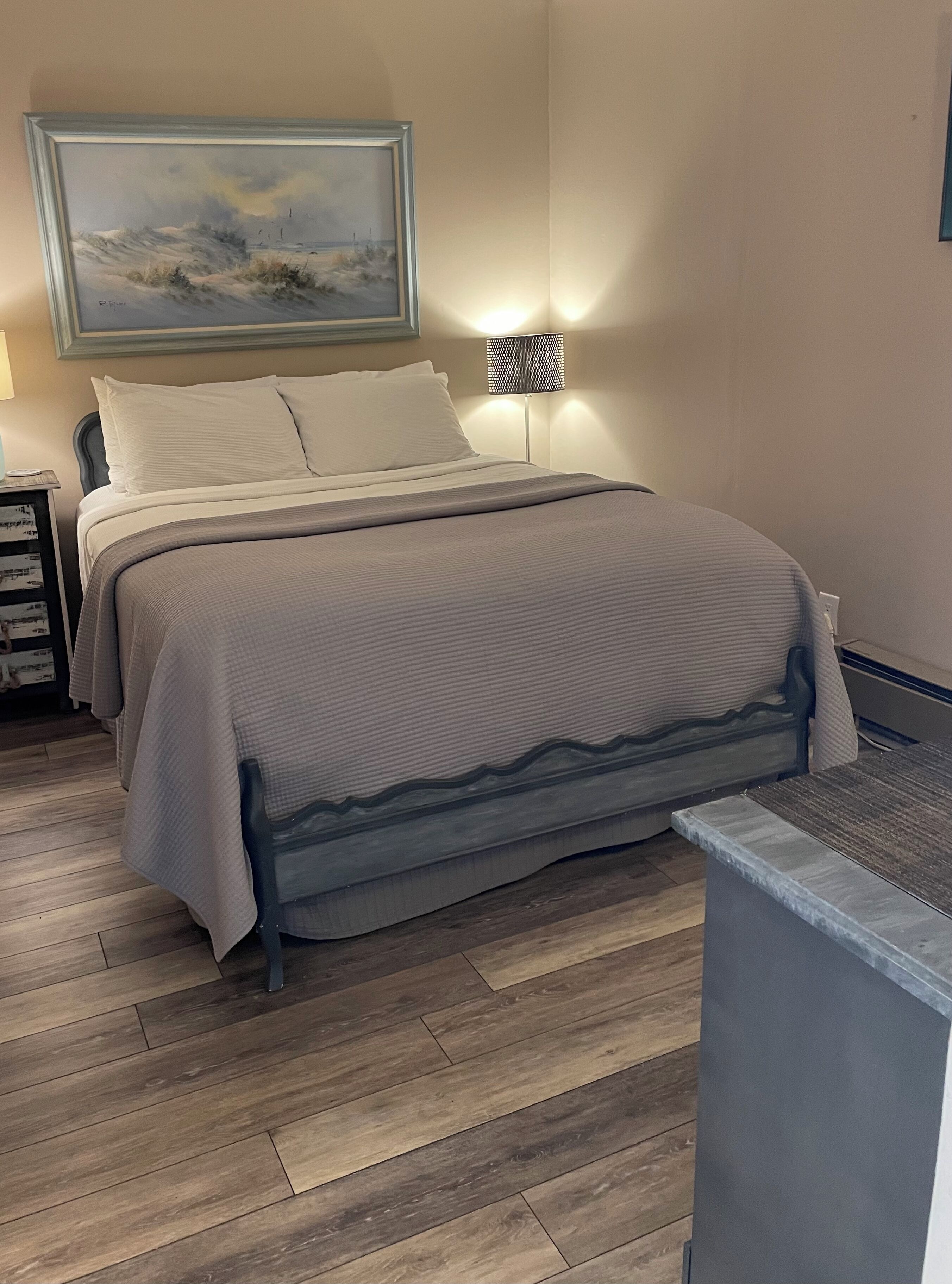 A neatly made bed with a gray blanket, a nightstand with a lamp, and a painting on the wall.