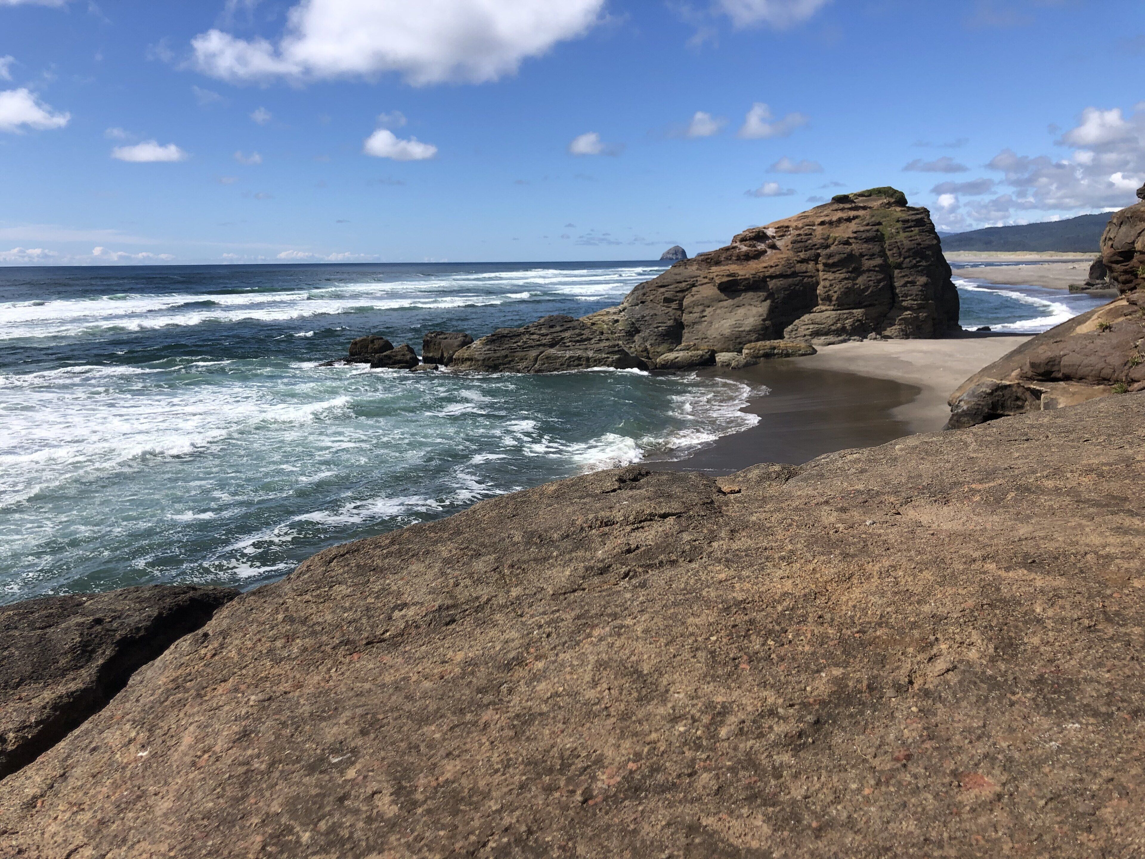 A rocky coastline with waves crashing onto the shore, a sandy beach, and a clear sky.