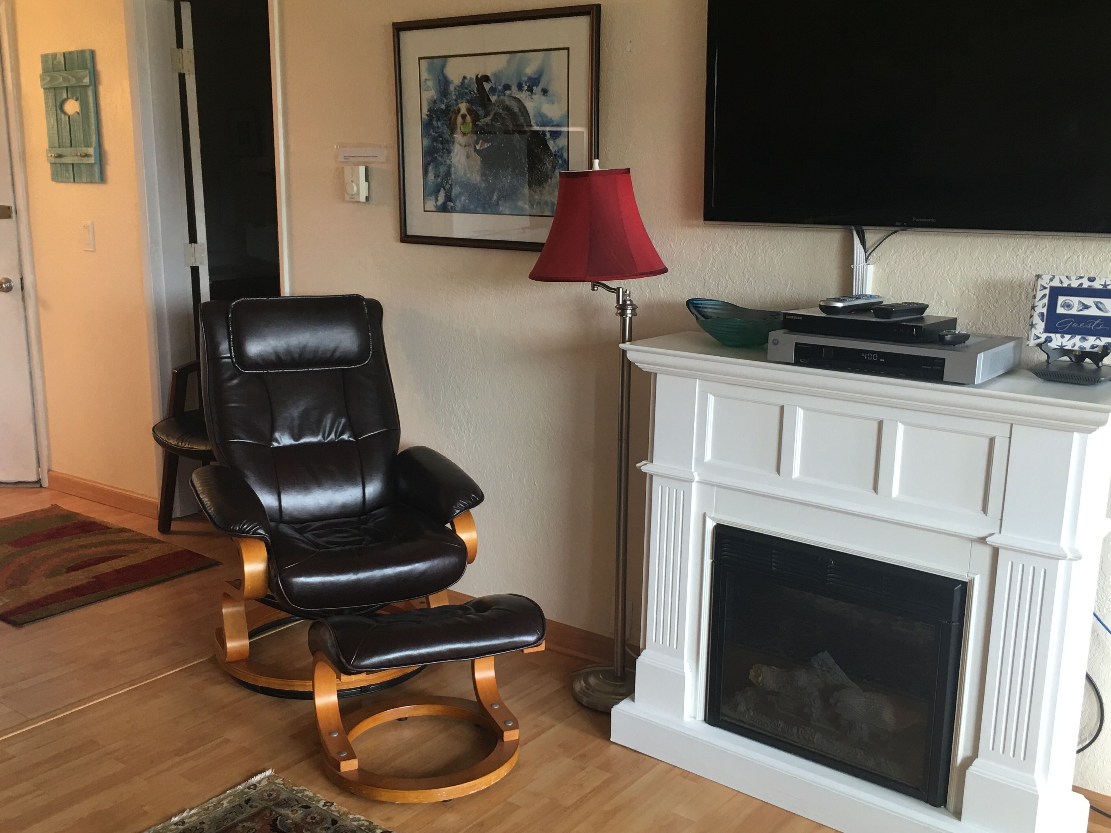 A living room with a black leather chair, a red floor lamp, a white fireplace, and a flat-screen TV mounted on the wall.