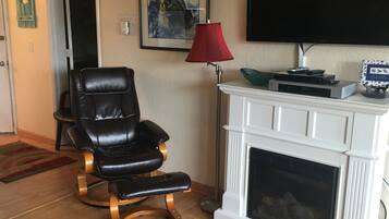 A living room with a black leather chair, a red floor lamp, a white fireplace, and a flat-screen TV mounted on the wall.