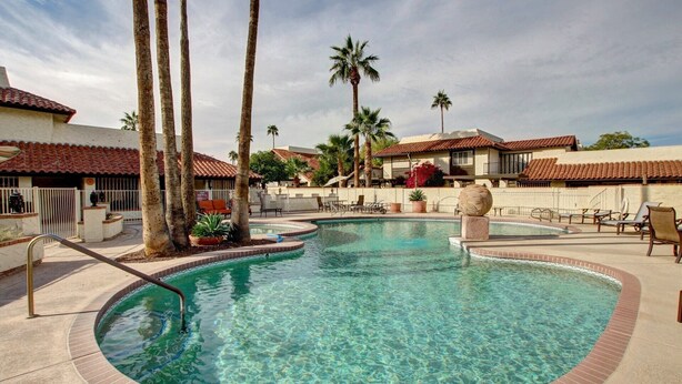 A clear swimming pool with a tiled deck, surrounded by palm trees and a building with a red-tiled roof.