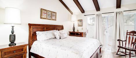 Un dormitorio con una cama de madera, una mesita de noche con lámpara, una mecedora y un ventanal con cortinas.