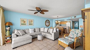 Ein Wohnzimmer mit einem Ecksofa, einem Rattan-Sessel, einem Deckenventilator und einer Küchenzeile im Hintergrund.