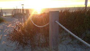 Un poteau en bois avec une barrière faite de cordes, sur une plage de sable, avec l’océan et le coucher de soleil en arrière-plan.