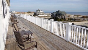 Une terrasse en bois avec des sièges d’extérieur, des rampes blanches et une vue sur la plage et l’océan.