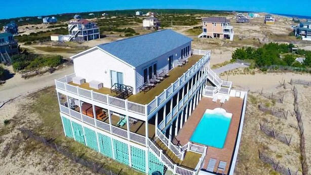 Une grande maison de deux étages située en bord de mer, dotée d’une piscine et de plusieurs balcons.