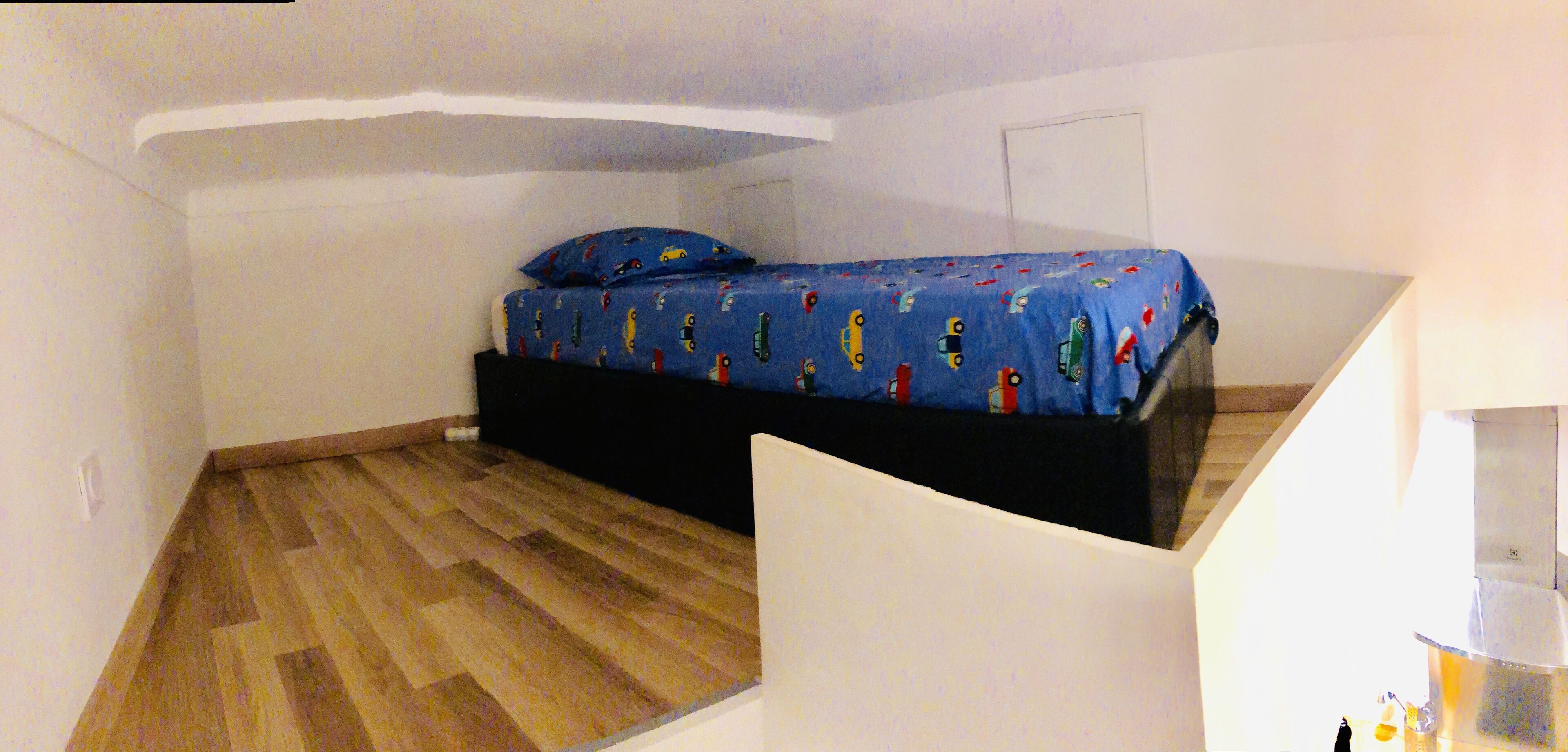 A bedroom with a bed on a black platform, a blue bedspread with car patterns, and a wooden floor.