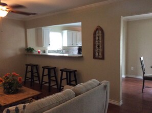 A living room with a sofa, a dining table with two stools, a kitchen area with white cabinets, and a decorative wall piece. - COZY, COMFY, CUTIE!  Book NOW for Fall!  2BD, 1.5 Bath, Free WIFI (Corpus Christi)