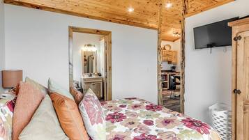 Een slaapkamer met een houten plafond, een bed met bloemenbeddengoed, een spiegel, een televisie en een keukenruimte zichtbaar door een open deur.