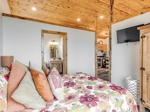 A bedroom with a wooden ceiling, a bed with floral bedding, a mirror, a TV, and a kitchen area visible through an open door. - Romantic Lakeview Treehouse with Jacuzzi Tub & Private Deck (Stigler)