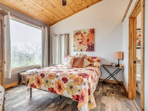 Un dormitorio con cama, ventana, un cuadro y mesita de noche.
