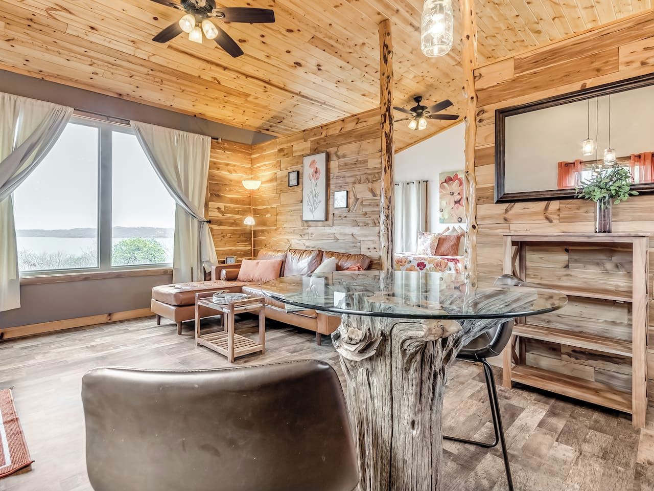 Een gezellig chaletinterieur met een eettafel met glazen blad, houten wanden, een bank en een groot raam.