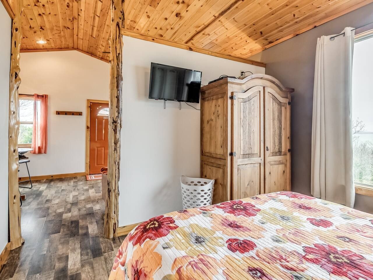 A bedroom with a wooden ceiling, a bed with a floral bedspread, a wardrobe, a television, and a window with curtains.