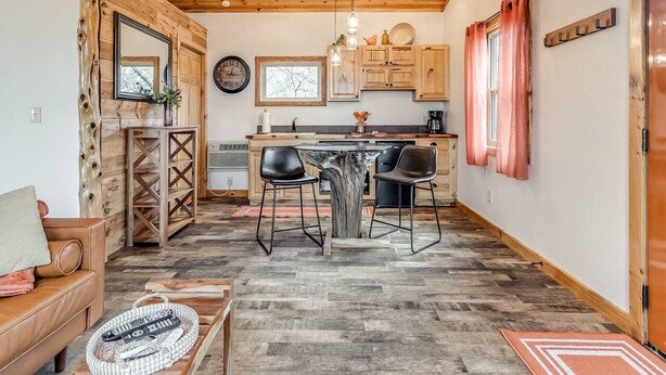 Un interior acogedor con techo de madera, un comedor con mesa y sillas centrales, una cocina con armarios y ventana, y un área de estar con sofá y mesa de centro.