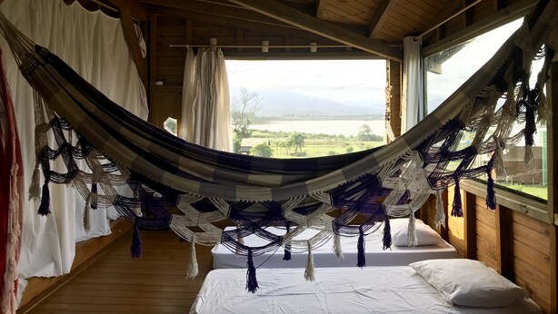A hammock suspended between wooden beams in a room with a bed and a view of a lake and mountains. - Casa Abacate (Imbituba)