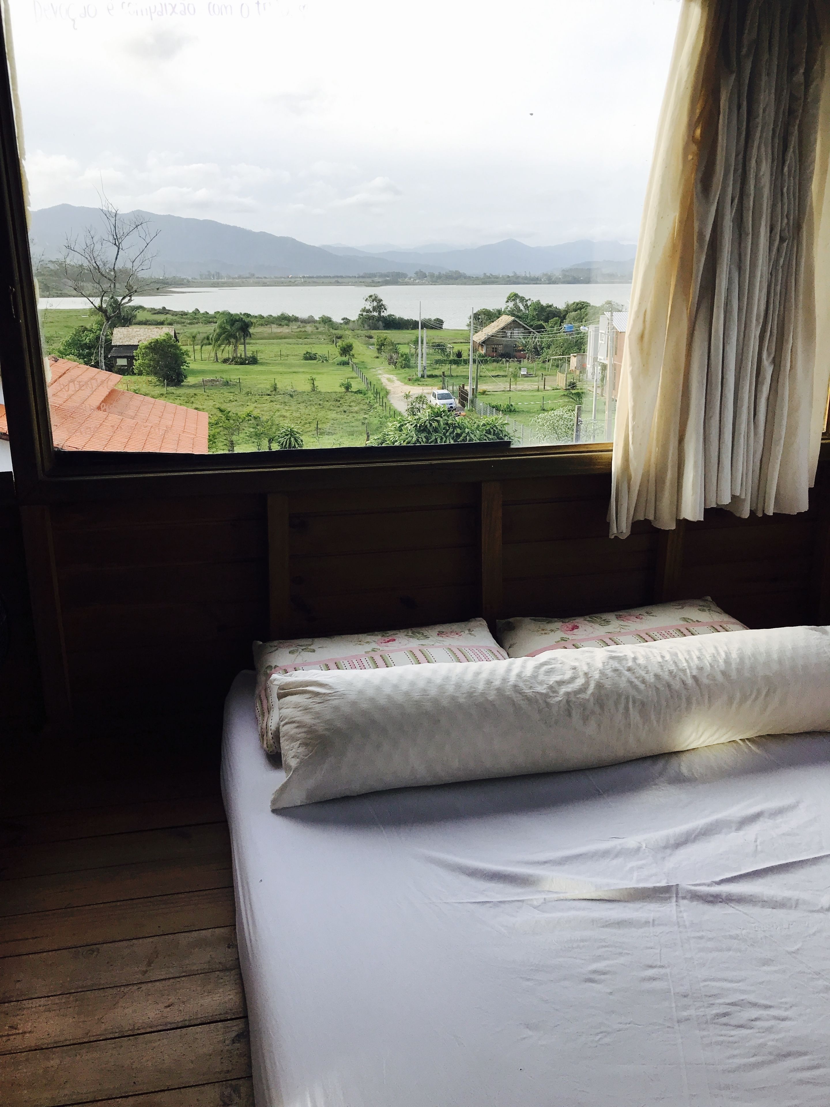 A room with a bed, wooden walls, and a window overlooking a landscape with a body of water and distant mountains.