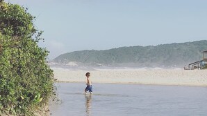 A beach scene with a child playing in shallow water, a sandy shore, and a forested hill in the background. - Casa Abacate (Imbituba)
